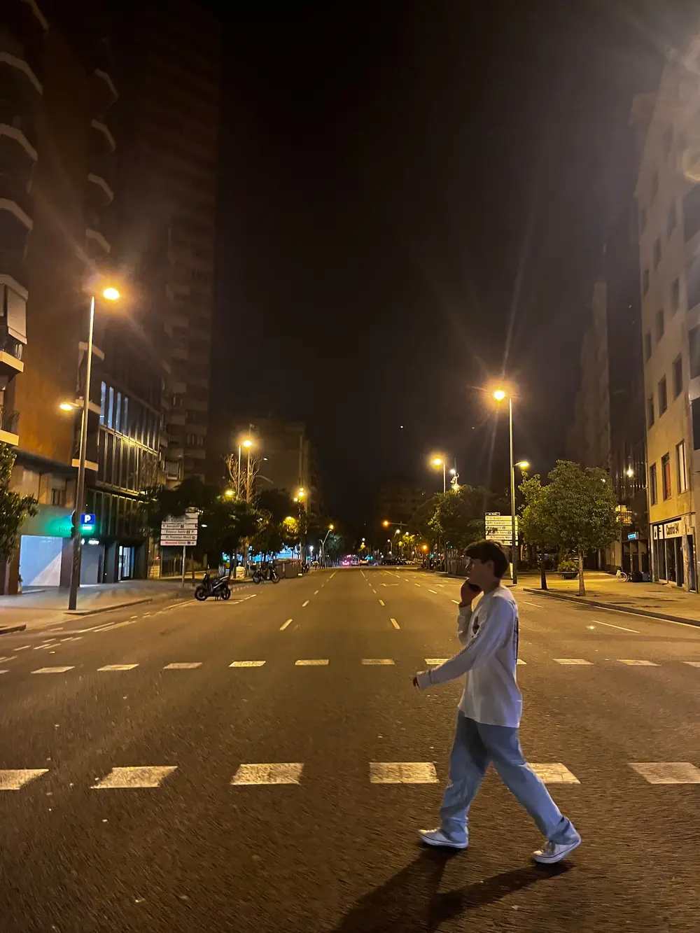 A person crossing a broad, nearly empty street under streetlights at night.
