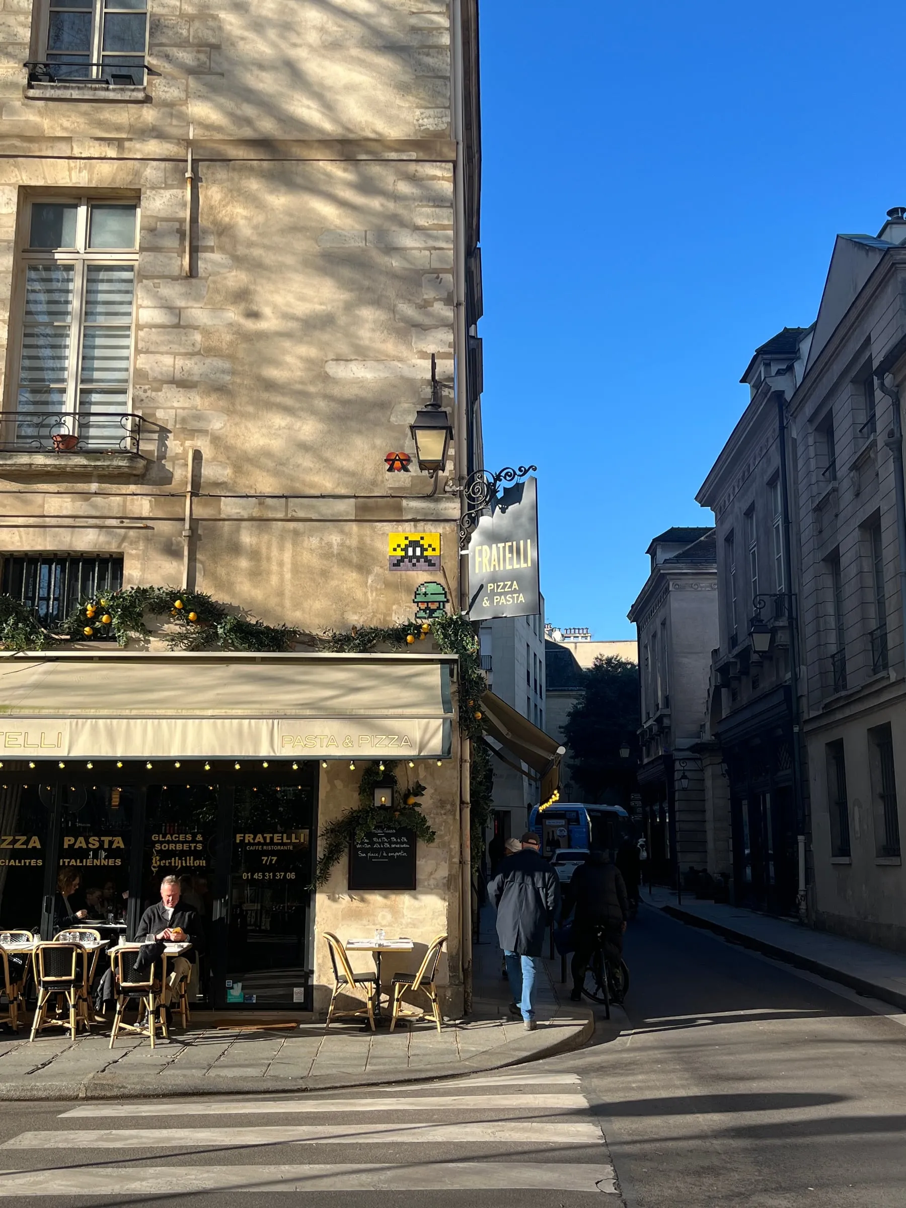 Sunlight and tree shadows across a corner cafe on a narrow Paris street.