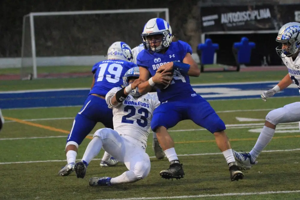 An American football player in blue holding the ball while a defender tackles him.