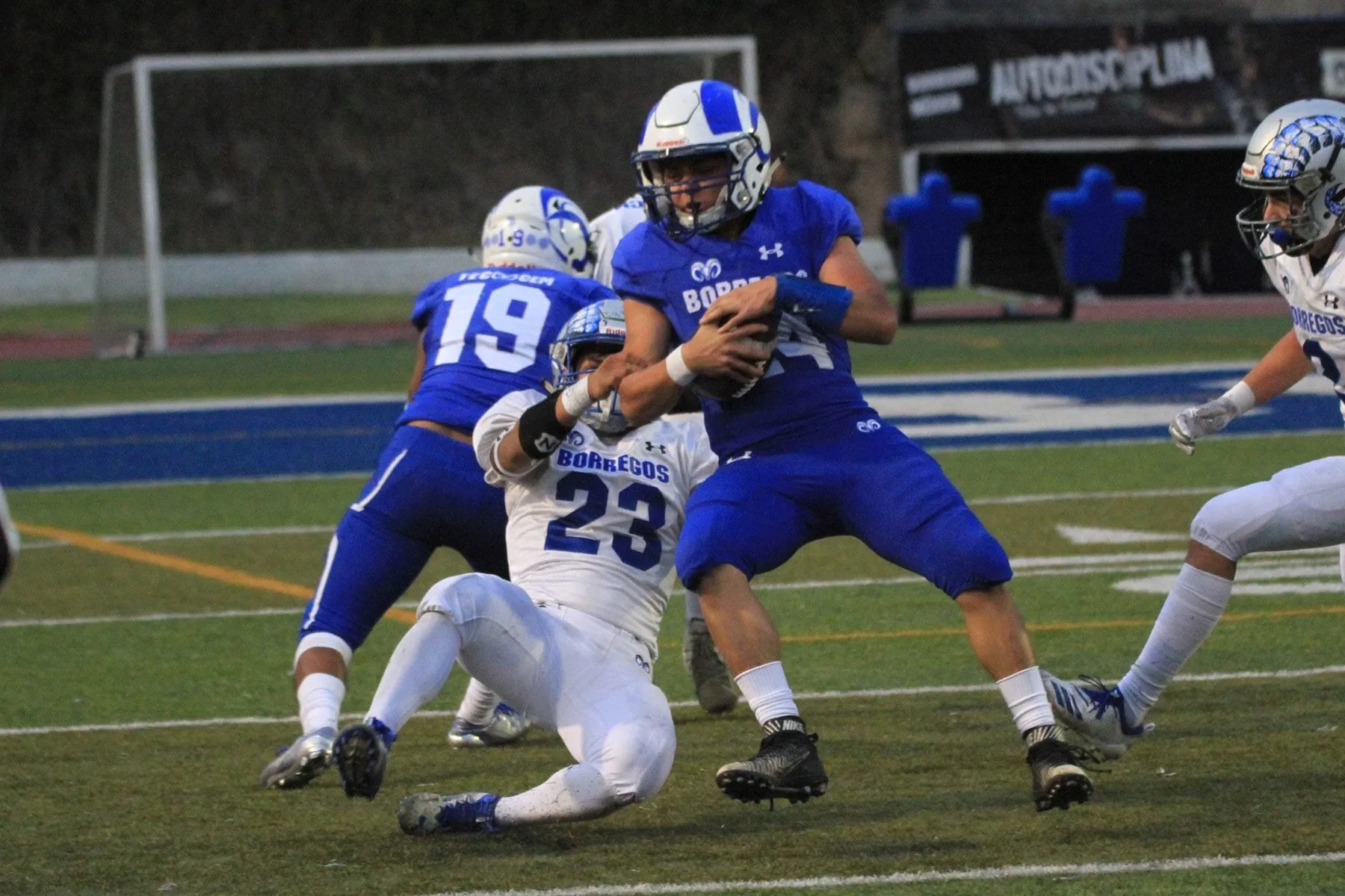 An American football player in blue holding the ball while a defender tackles him.