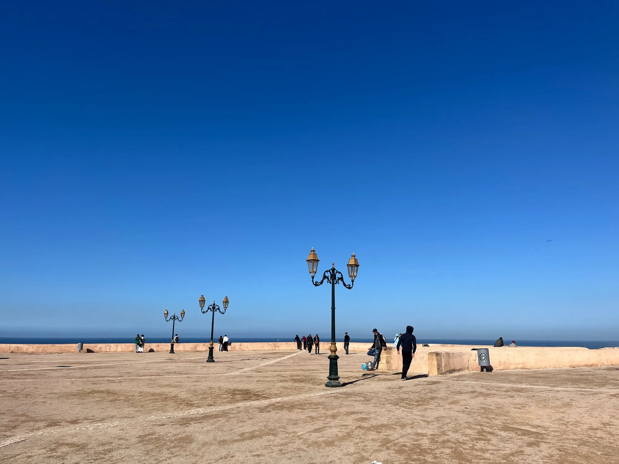 People and ornate lampposts on a broad seaside promenade under a clear blue sky.