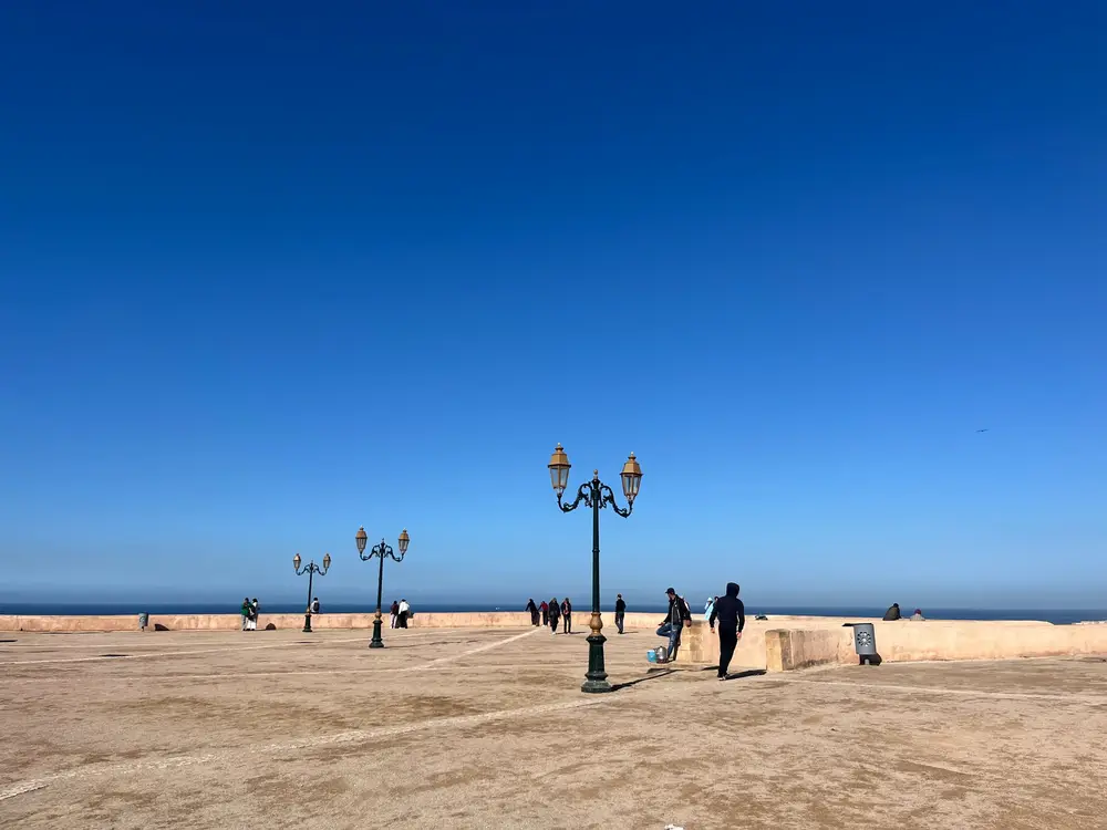 People and ornate lampposts on a broad seaside promenade under a clear blue sky.
