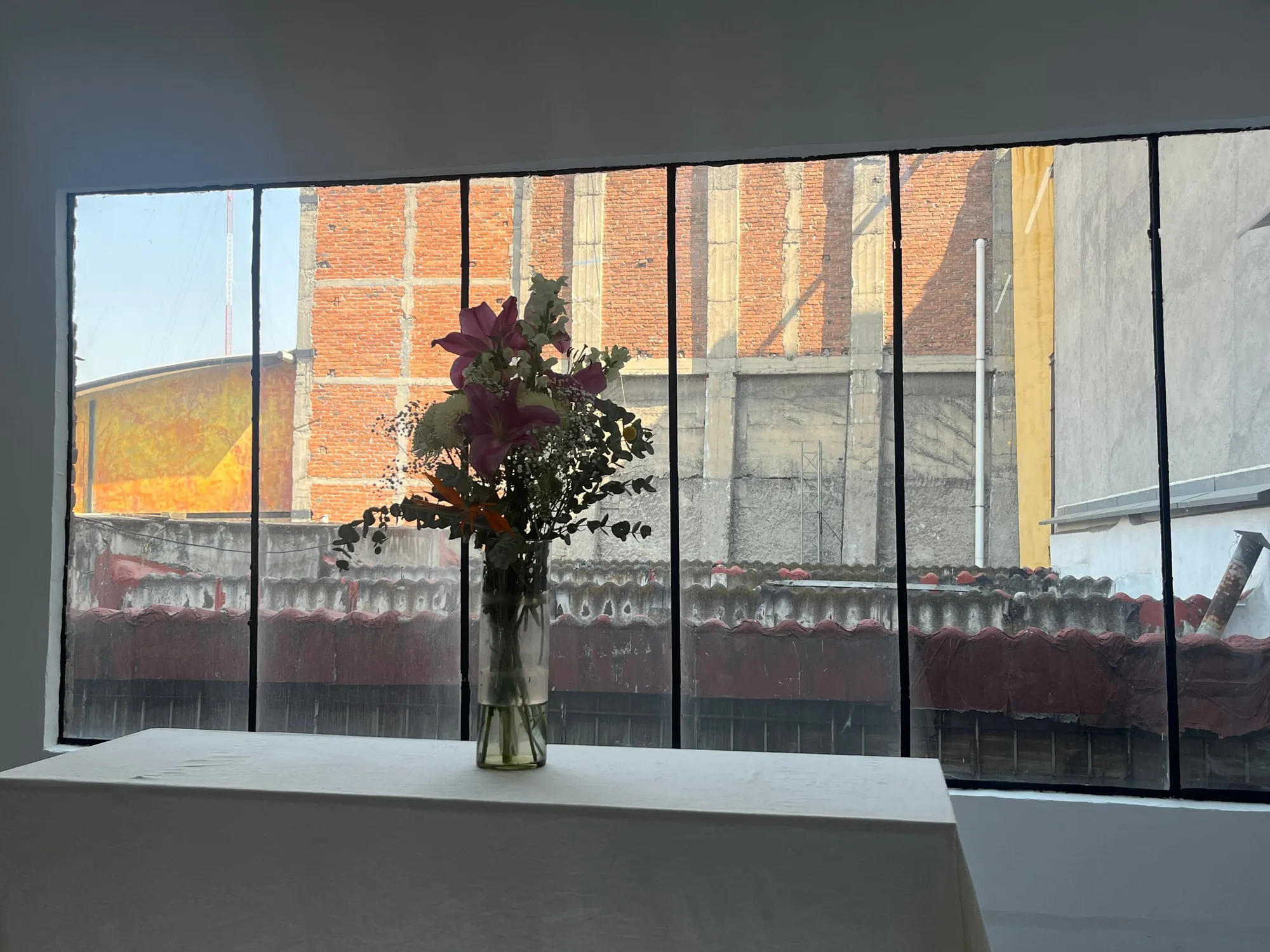 A vase of flowers on a white table in front of large windows.