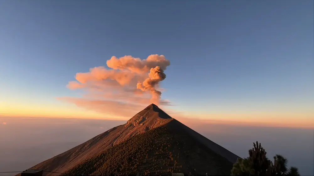 A plume rising from a steep volcano against an orange and blue dawn sky.