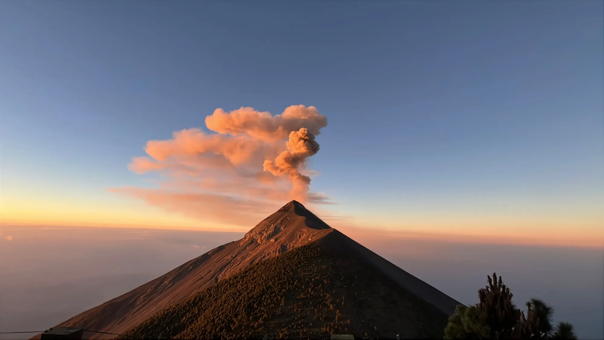 A plume rising from a steep volcano against an orange and blue dawn sky.