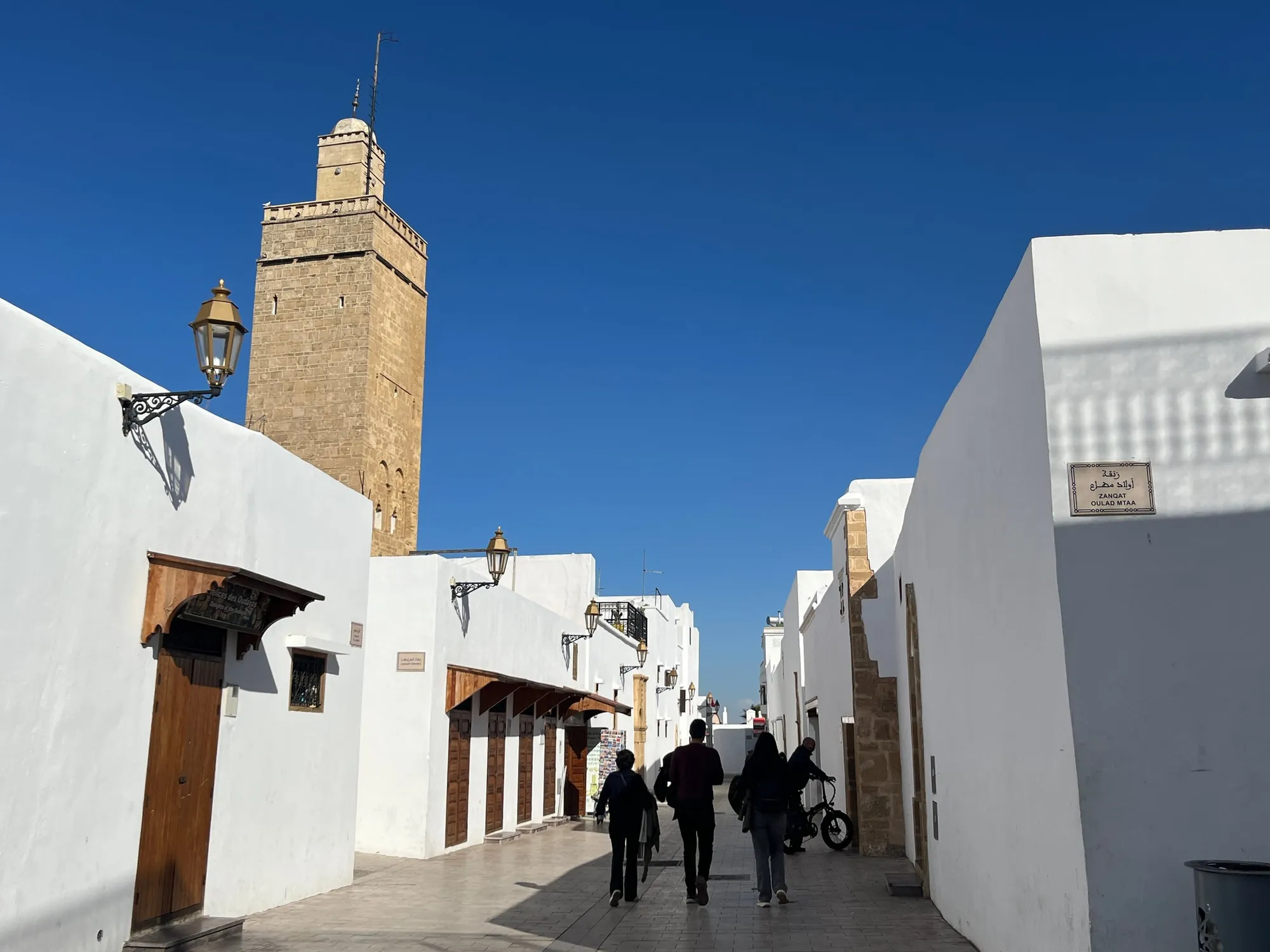 Pedestrians walking between white walls beneath a tall stone minaret.