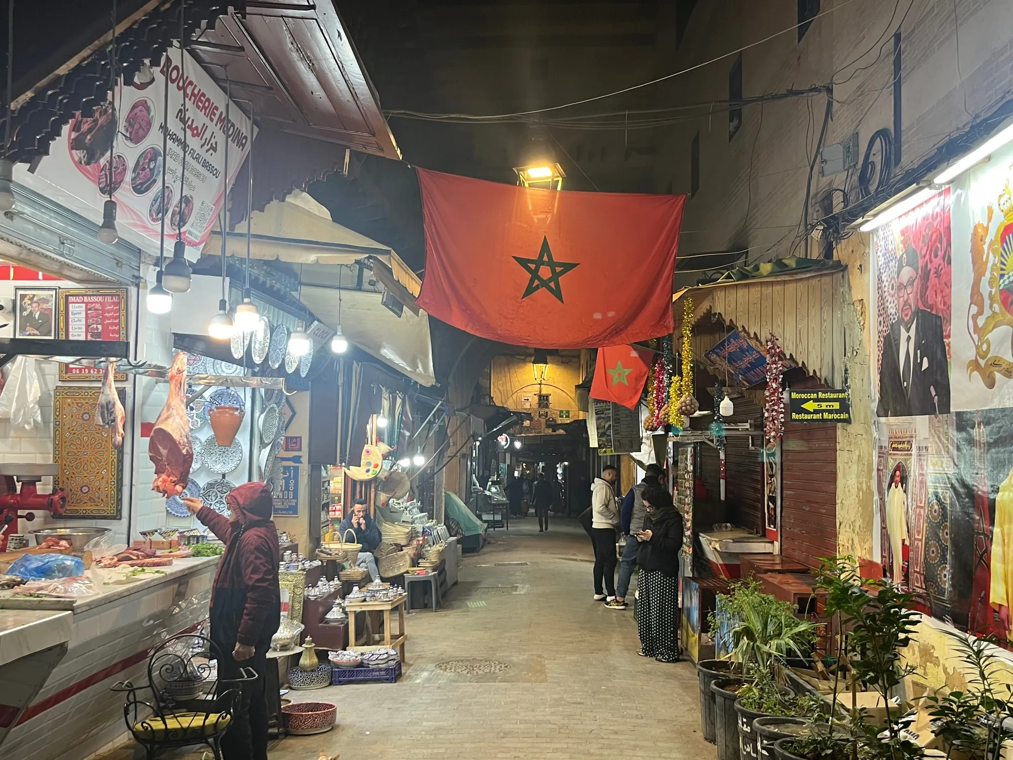 A narrow market passage lined with stalls beneath a Moroccan flag.