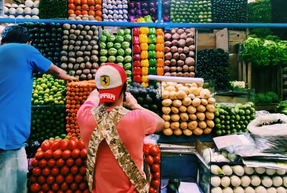 A person in a red cap and pink shirt in front of neatly stacked fruit at a market stall.
