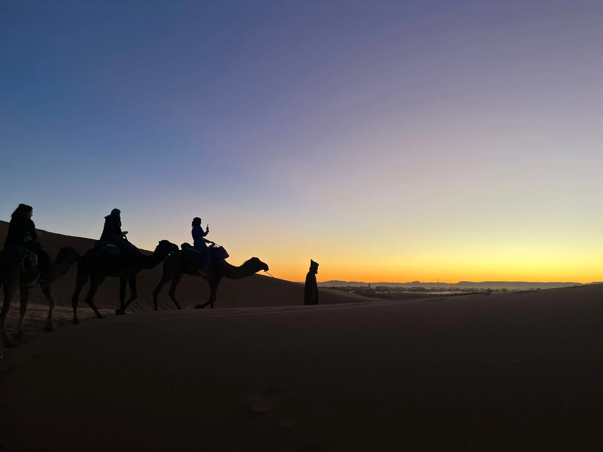 Silhouettes of riders on camels crossing sand beneath a glowing sunset sky.