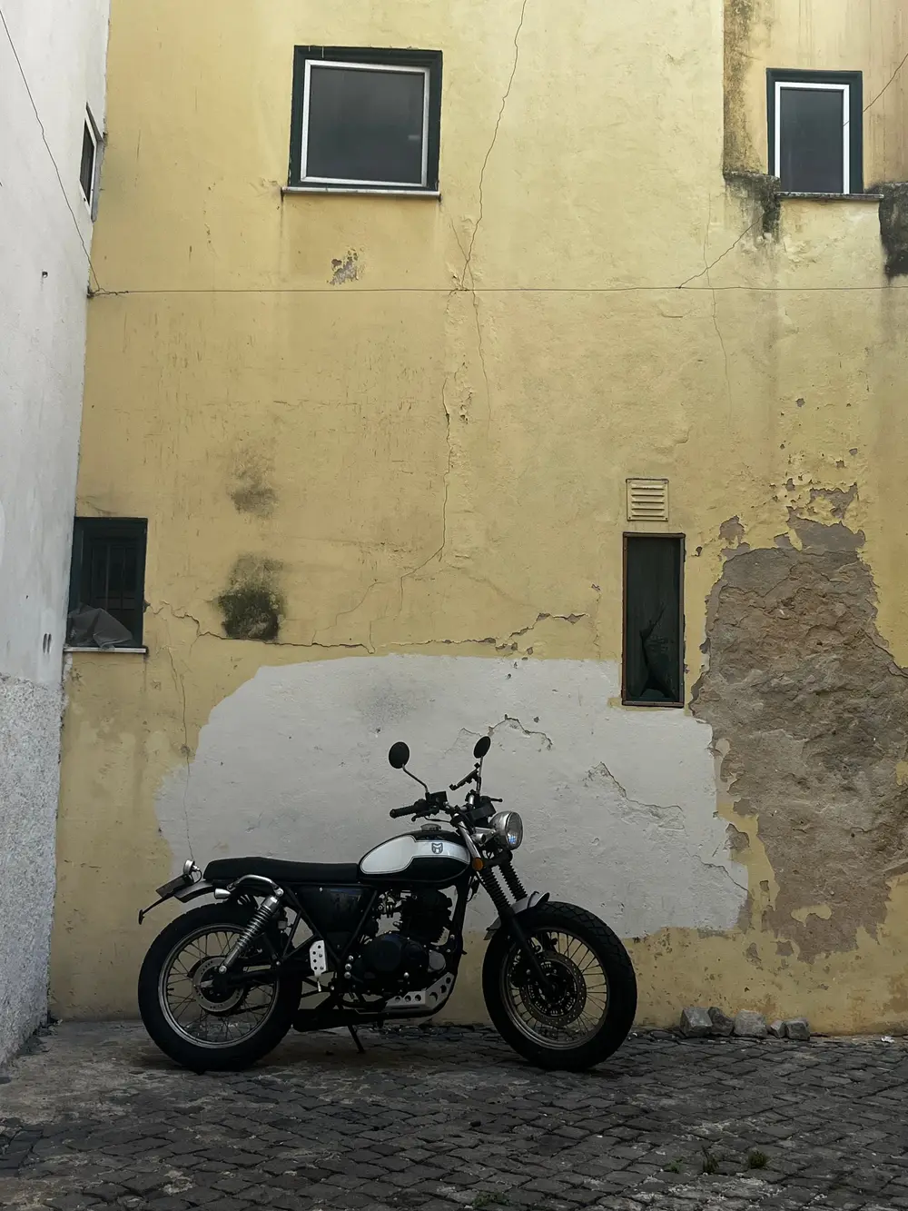 A black motorcycle parked against a weathered yellow wall with small windows.
