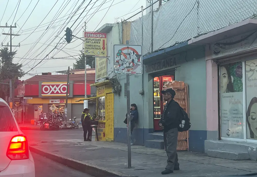 People waiting by an intersection beneath overhead cables and storefront signs.