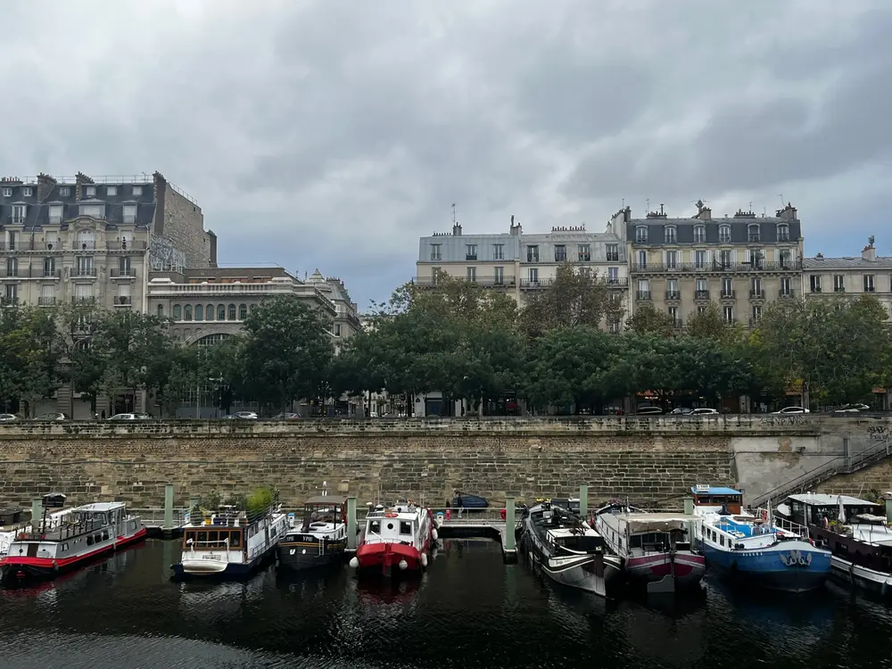 Boats moored along a tree-lined canal beneath apartment buildings and a cloudy sky.
